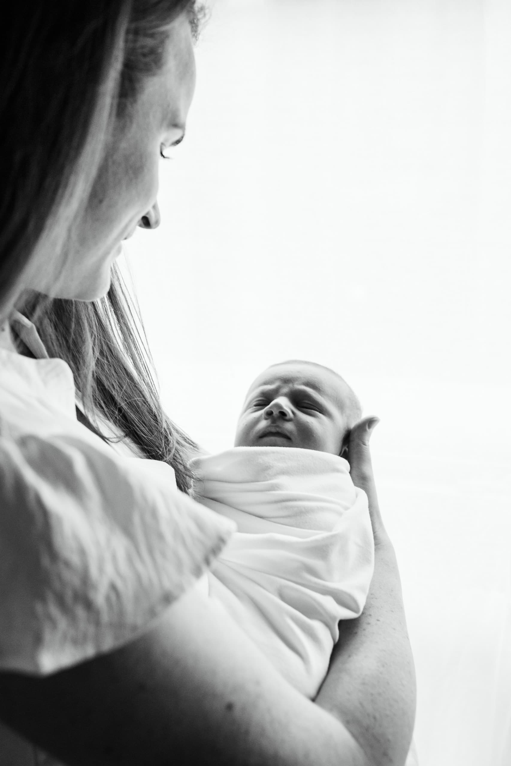 A mother gazing at her swaddled newborn in soft window light, black and white